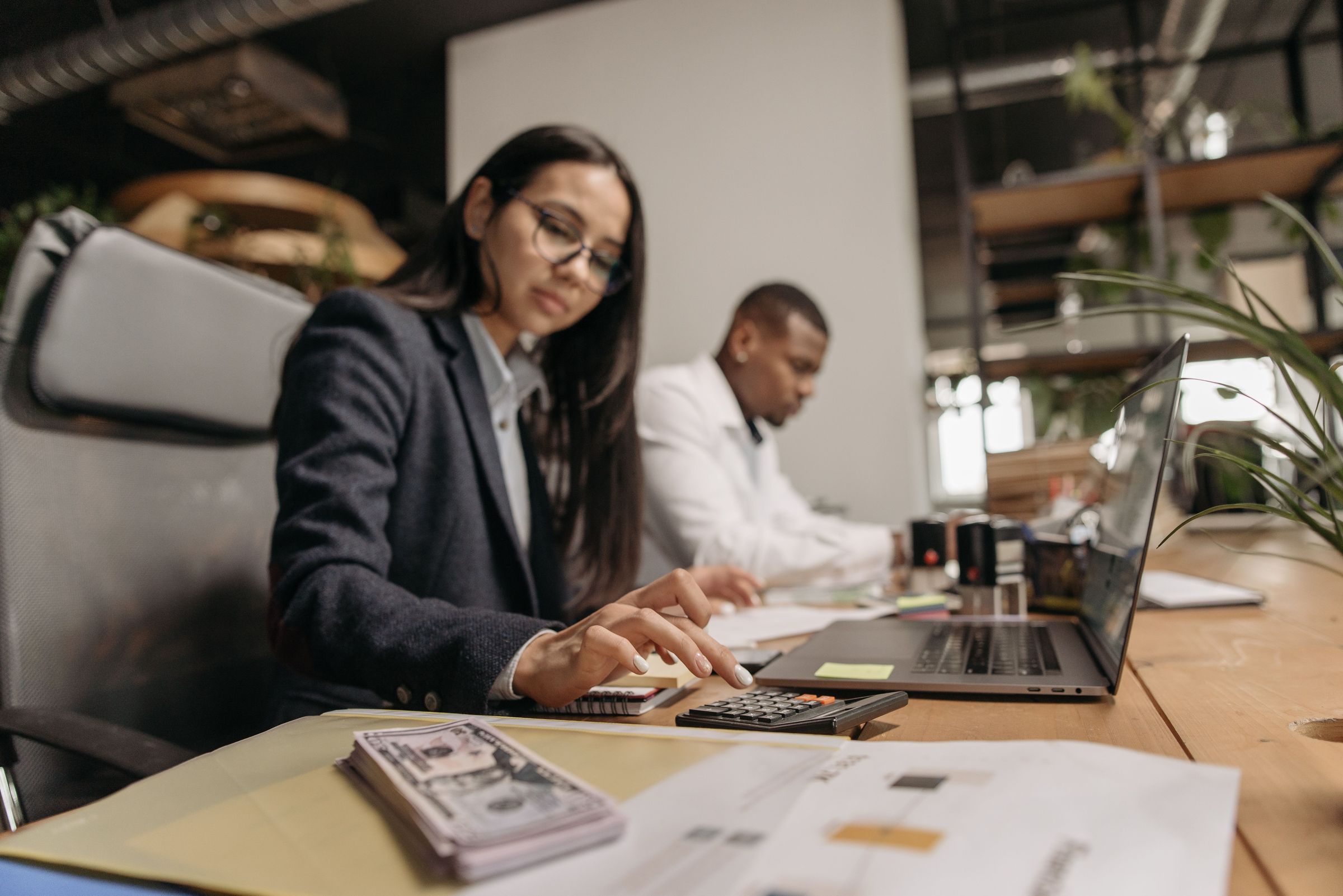 Two colleagues at a shared desk in a bright office, one working a calculator and laptop while the other works behind her