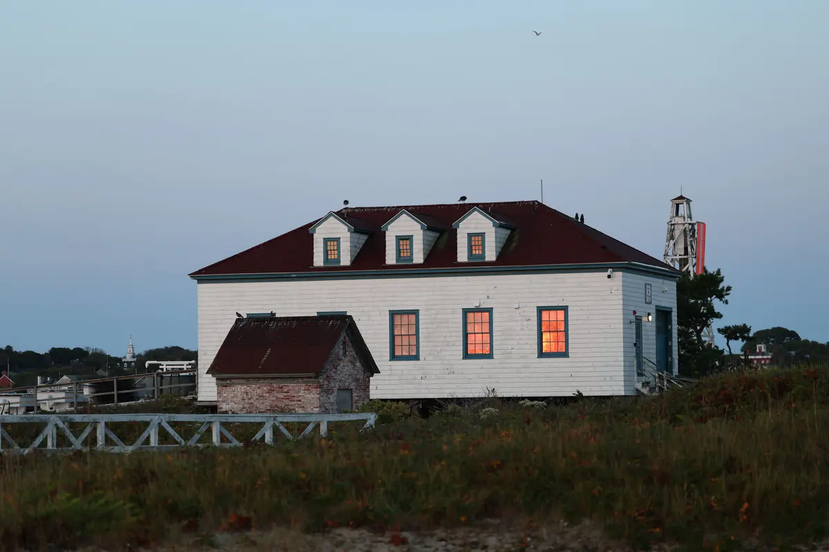 A white shingled coast guard station with a red roof and lit windows at dusk, with a lighthouse beside it