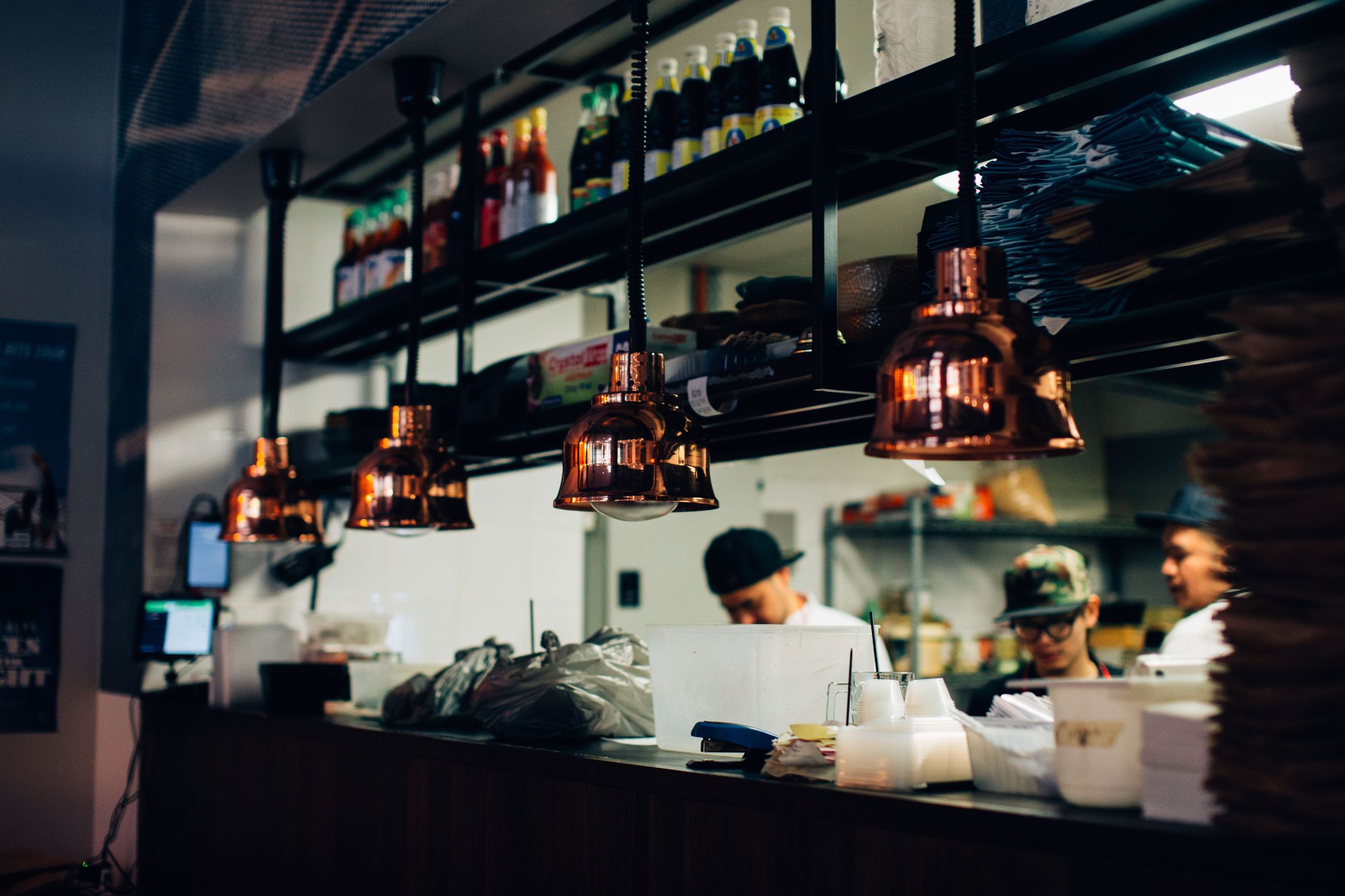 Cooks at the pass of an open restaurant kitchen under copper heat lamps, with the bar and dining room beyond