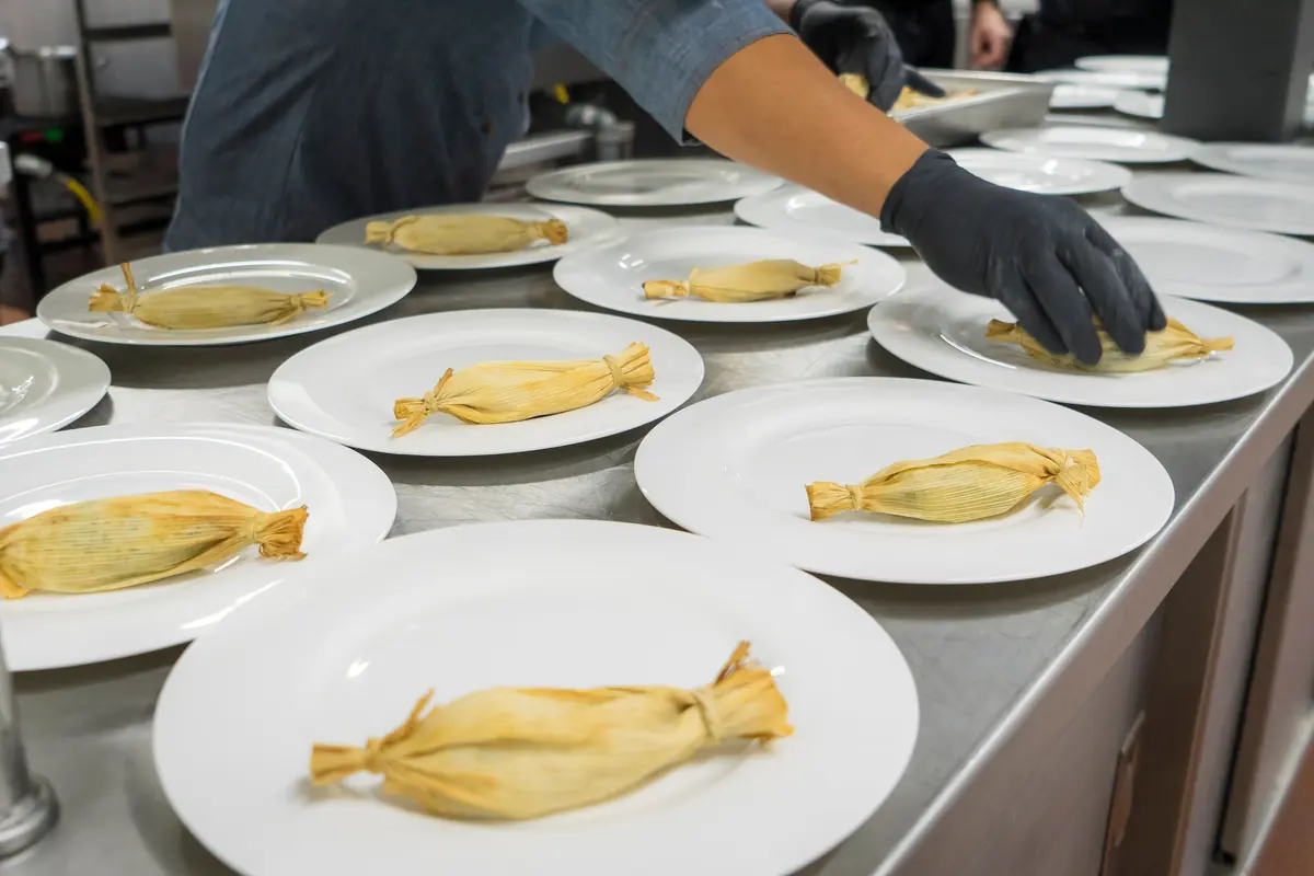 A gloved hand setting a wrapped tamale on each of a row of white plates on a steel pass