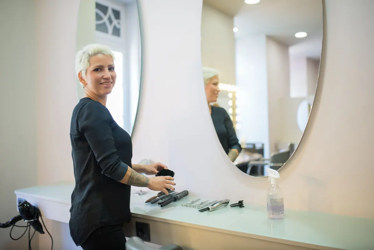 A salon owner in a black top standing beside two round mirrors and smiling at the camera