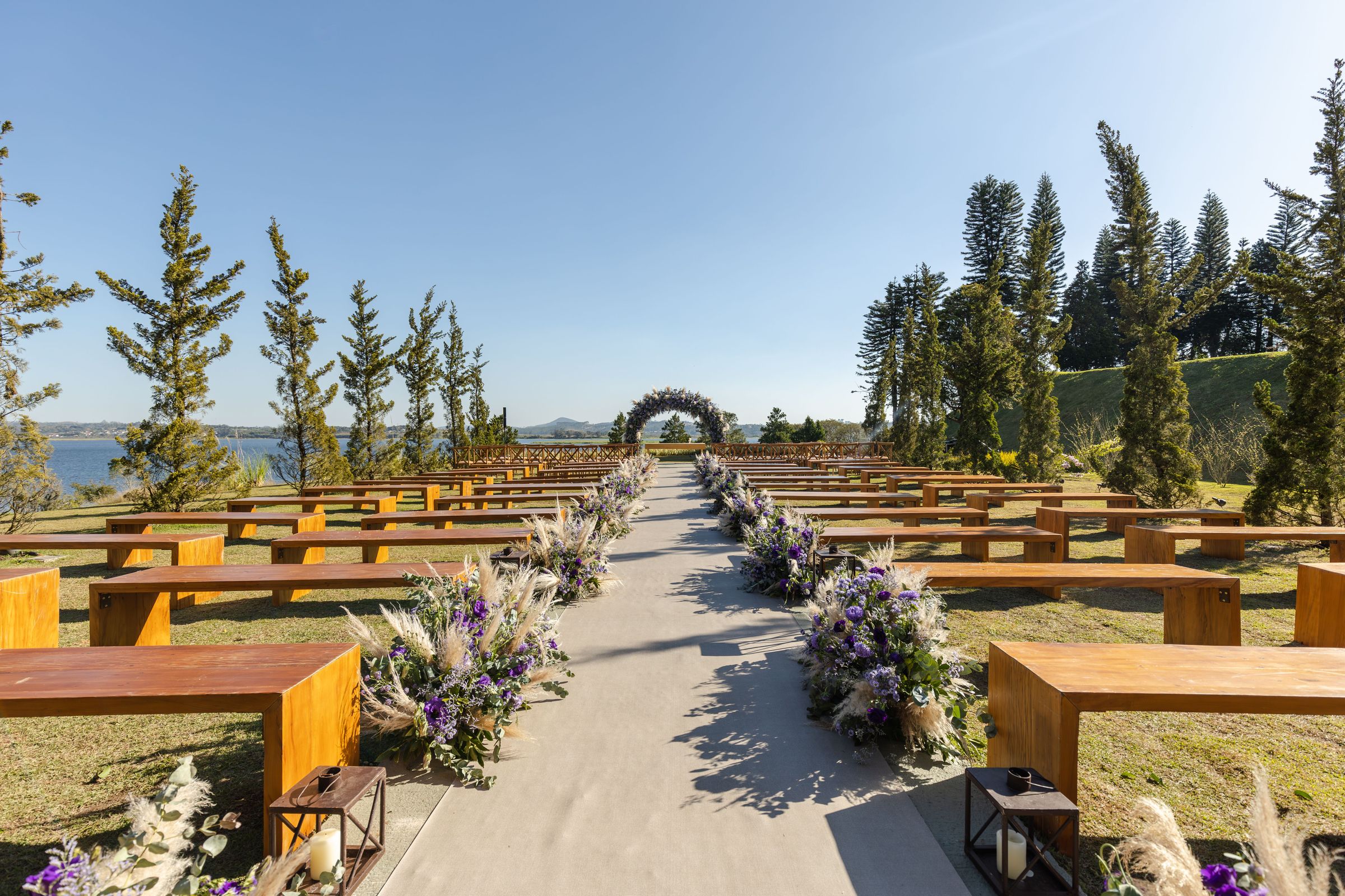 A lakeside wedding venue with rows of wooden benches, a flower-lined aisle and a floral arch under a clear blue sky