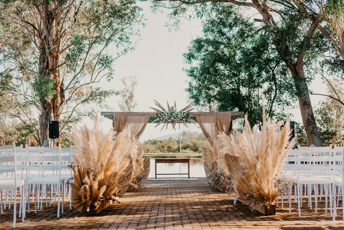 An outdoor ceremony set up with white chairs, pampas grass and a draped wooden arch under tall trees