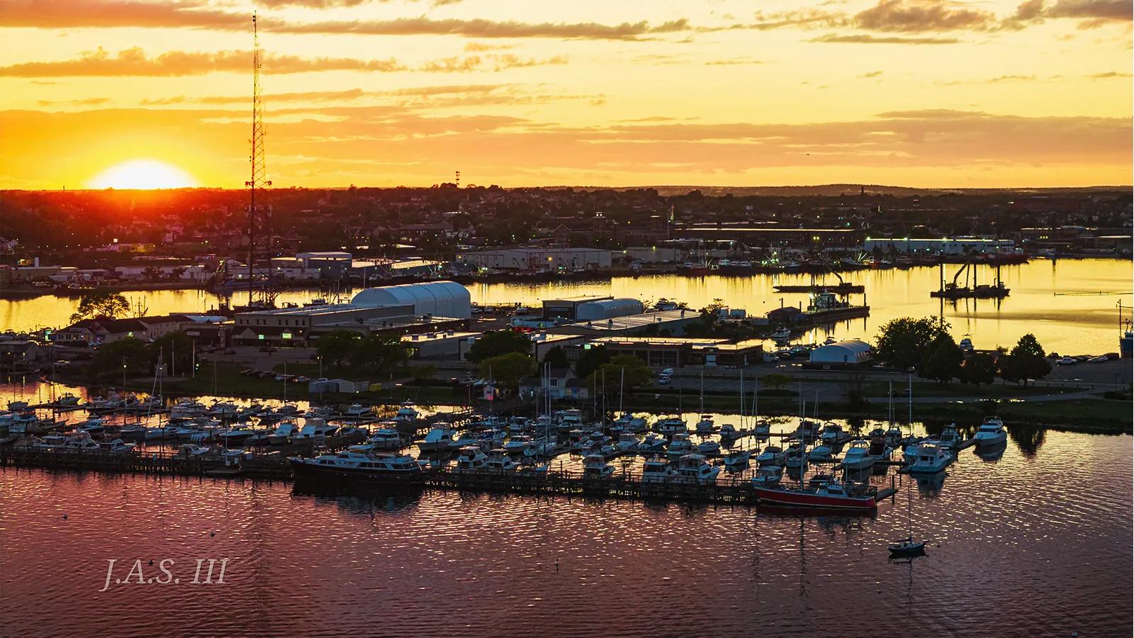 Sunset over a South Coast harbor, with a boatyard, work sheds and a marina full of boats on the near shore