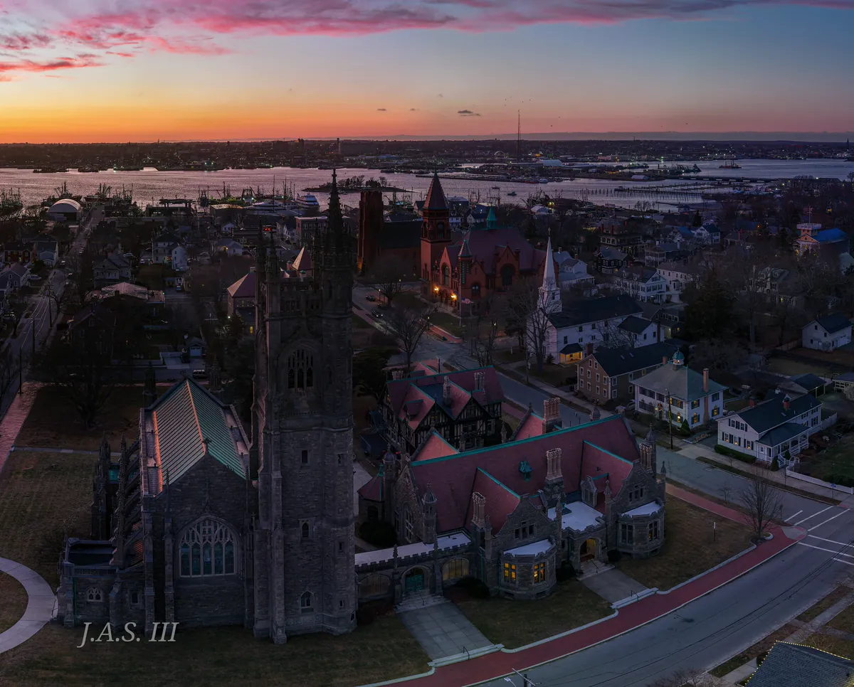 An aerial view at dusk of a stone church and old neighborhood buildings, with the harbor and a pink sky behind