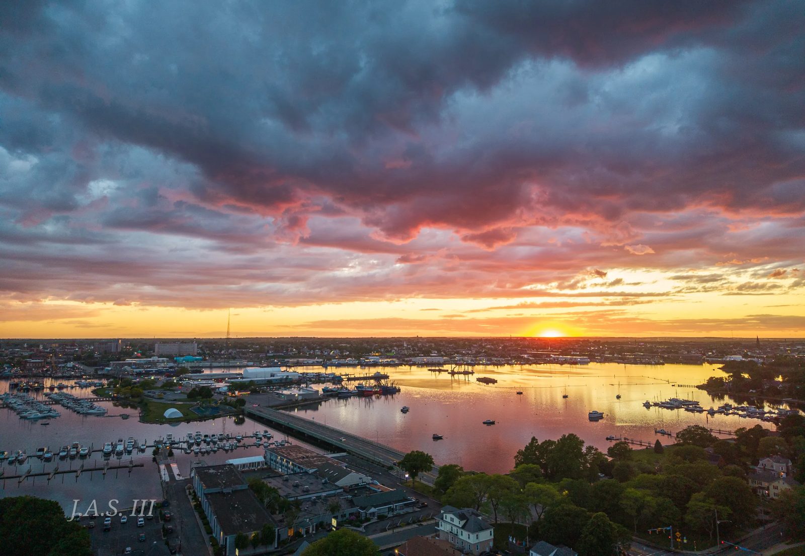 A harbor from above at sunset under heavy clouds, with marinas on both shores and a bridge across the water