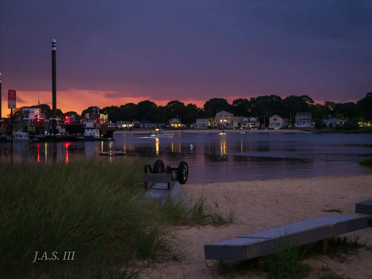 A harbor shore at dusk, with moored boats and the lights of the far bank on the water